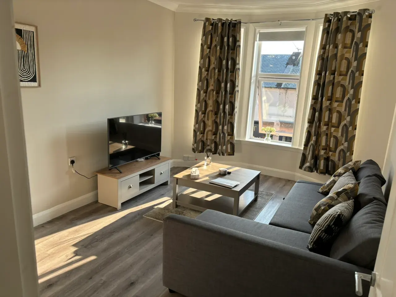 Bright living room with grey sofa, TV and a big bay window onto Ayr Street